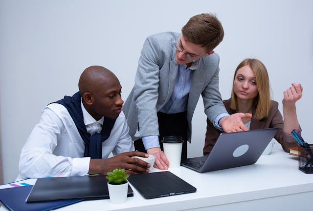 three people in work clothes at a desk discussing in front of an open laptop
