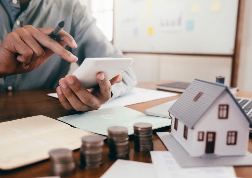 person holding a calculator with coins and home figurine in front of them